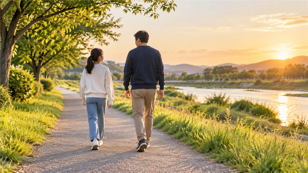 明るい夕方の川沿いで、少し間をあけて並んで歩く日本人の中学生と父親の後ろ姿
